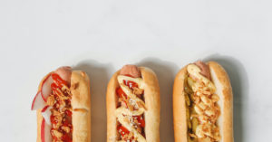 An aerial view of the top half of three hot dogs, each in a classic hot dog bun, aligned vertically: One with ketchup, sliced apples, and an indiscriminate crunchy topping; one with ketchup, mustard, and an indiscriminate crunchy topping; and one with ketchup, mustard, sliced pickles, and an indiscriminate crunchy topping. Each hot dog has a drop shadow to its right.
