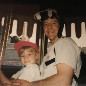Happy Father's Day: A father, wearing a white t-shirt and a smile, sits on a carousel with his daughter, a smiling blonde girl wearing a pink cap.