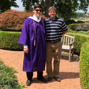 Happy Father's Day: Franklin, wearing purple graduation garb, stand alongside his father, wearing a striped shirt and khaki pants, in a sun-splashed patio pathway/garden. 