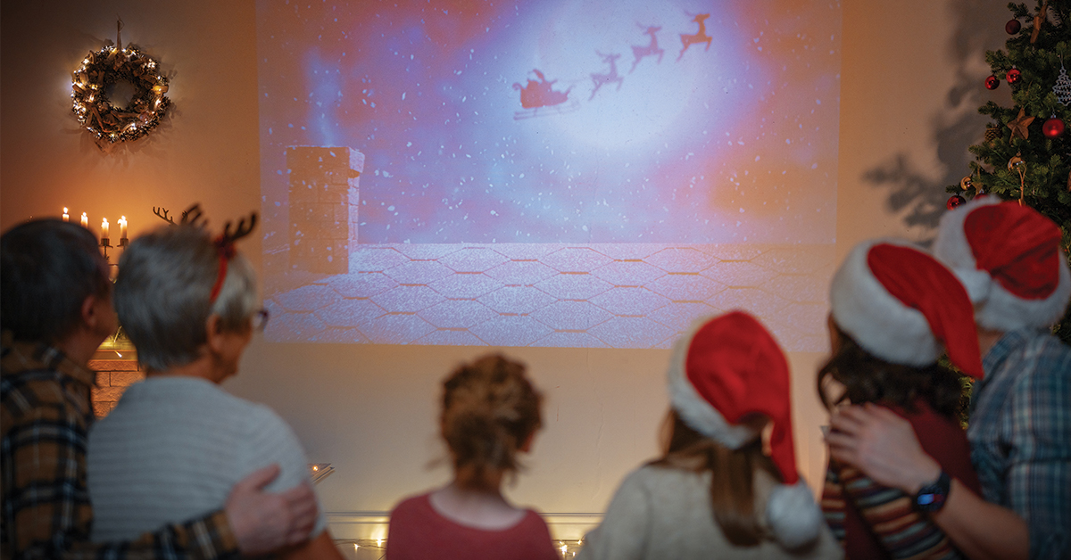 TQS takes on holiday movies: A view from behind a family of six who are seated on a couch, wearing festive holiday attire, and watching a projection of a holiday-themed movie depicting an animated Santa Claus and his reindeer flying away from a rooftop at night on Christmas Eve.