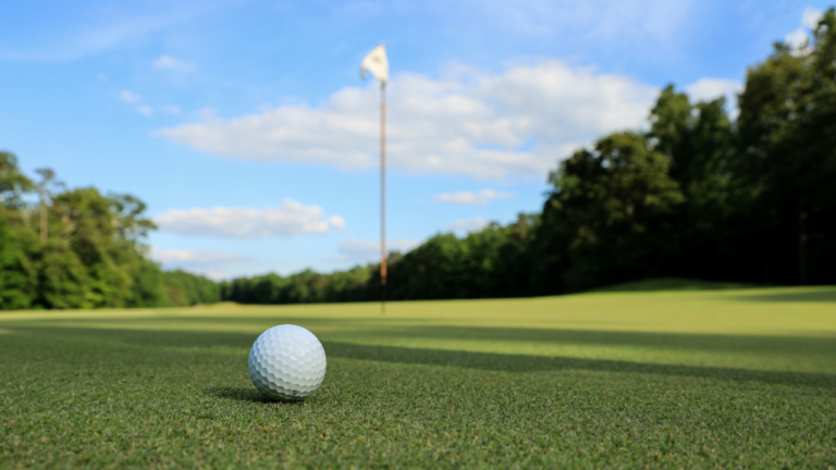 TQS takes on golf: A golf ball sits on the green with a flag flying in the background on a sunny day with blue skies and white clouds.