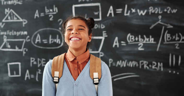 An adorable grade-school girl smiles brightly in a powder blue cardigan, orange collared shirt, and backpack. A chalkboard with mathematical equations is in the background.