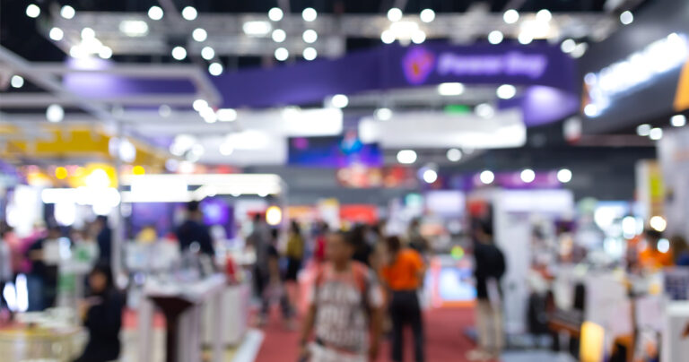 a blurry image of a convention floor with trade show booths and people walking about
